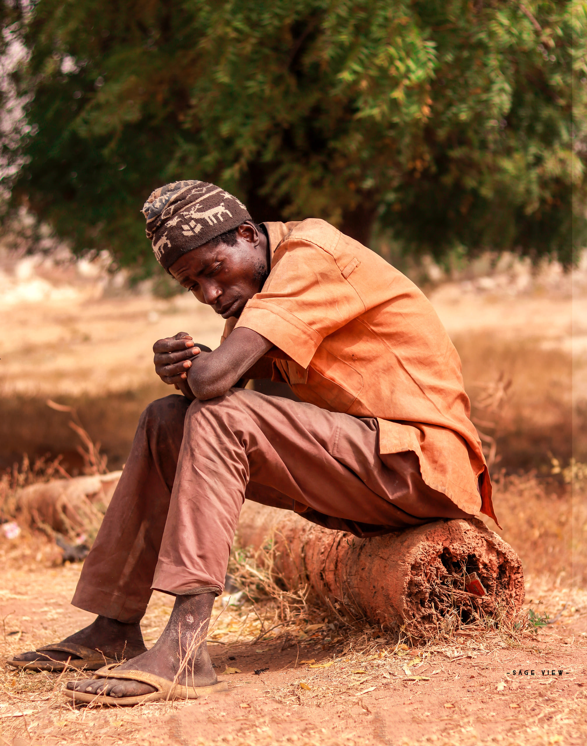 A boy in an orange shirt seated on a fallen log in dry rural scrub.